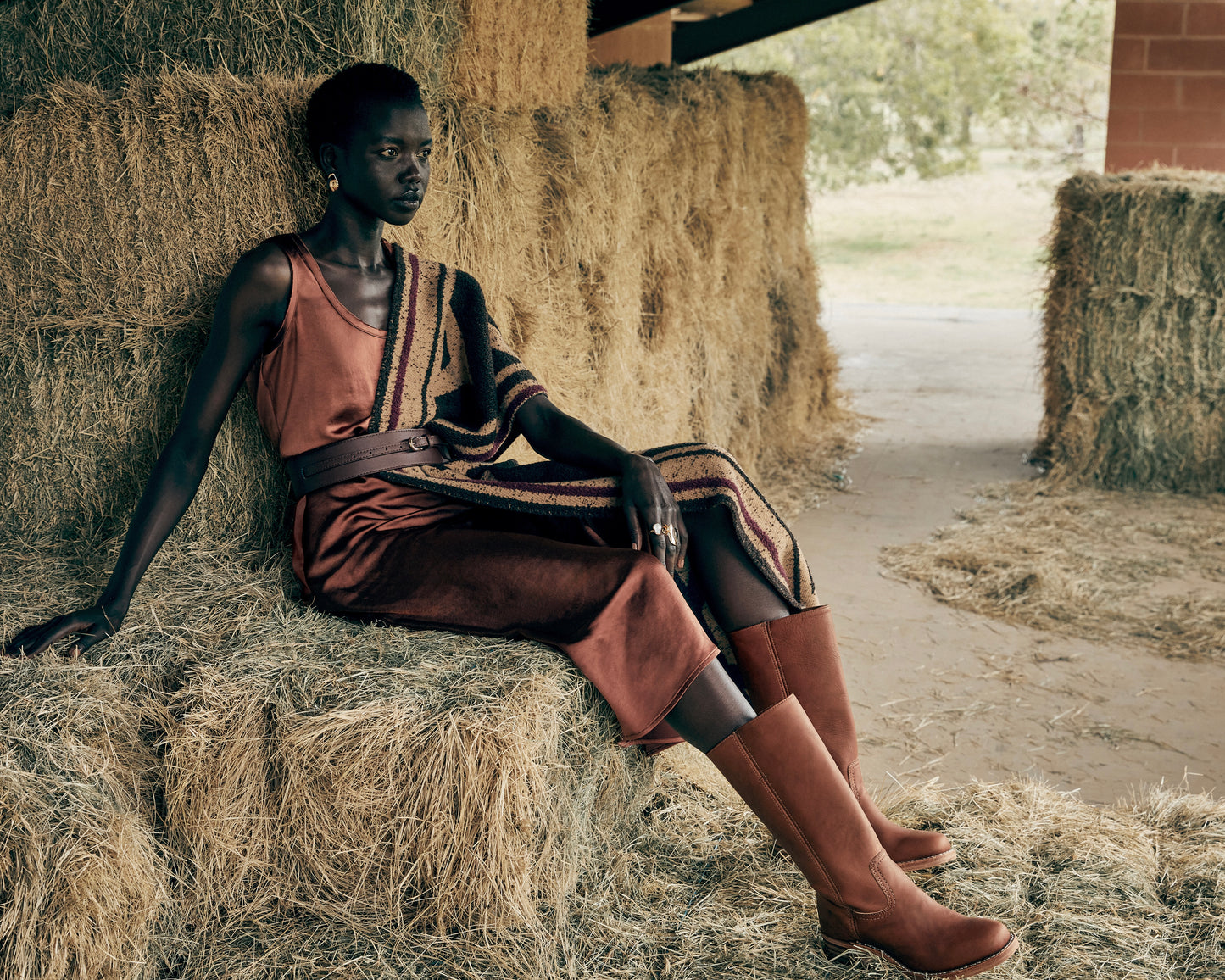 Woman in a rust colored slip dress sitting on hay bales in a rustic setting and wearing the Frye Campus 14L Boots in the color Saddle.