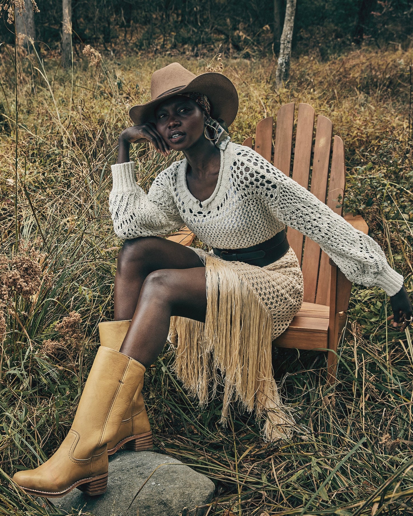 Woman sitting on a wooden chair in a natural setting wearing a white crochet sweater, a wide-brimmed hat, and the Frye Campus 14L Boots in the color Banana.