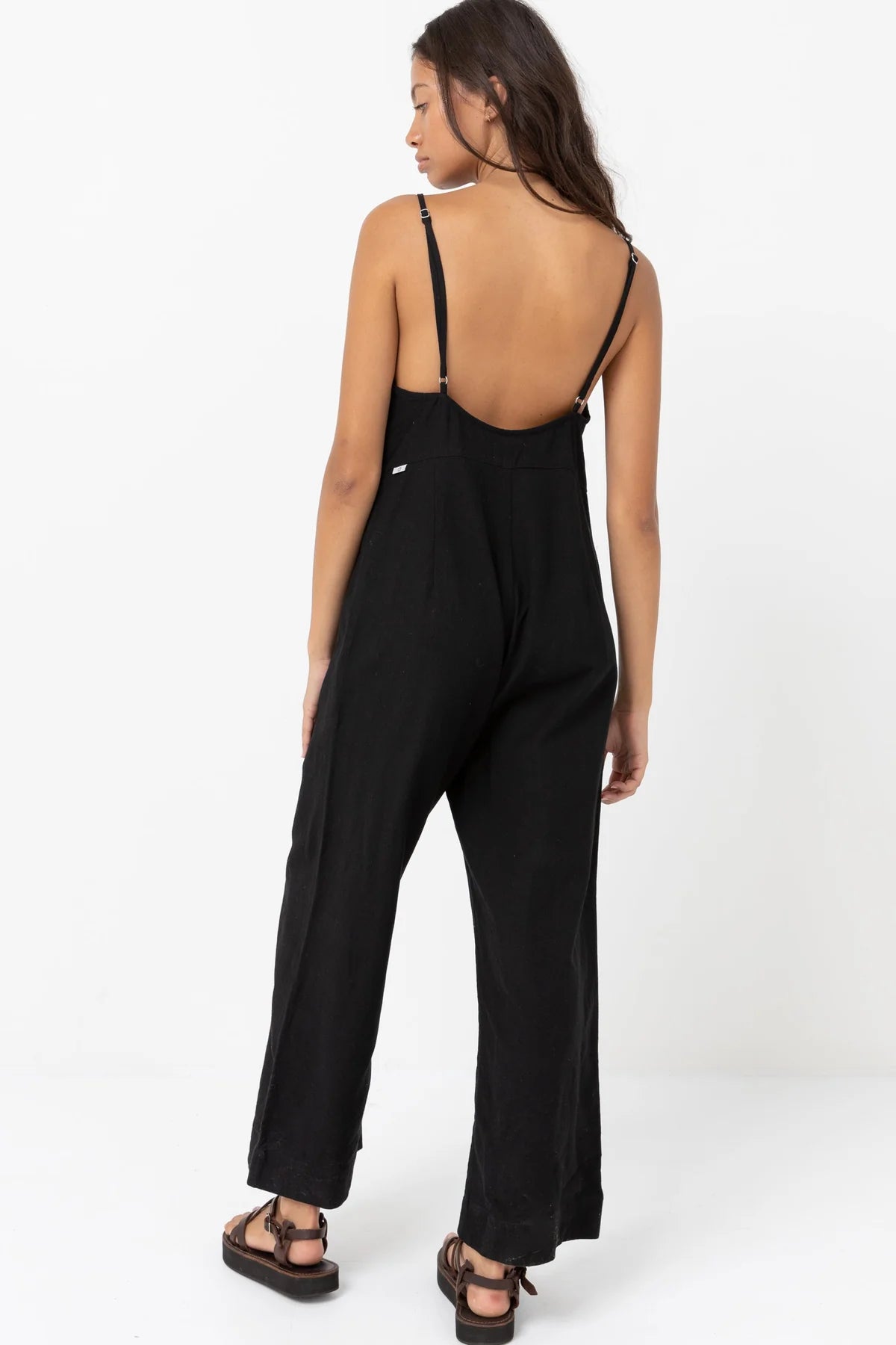 Back view of woman wearing a black, linen blend wide leg jumpsuit with adjustable straps
