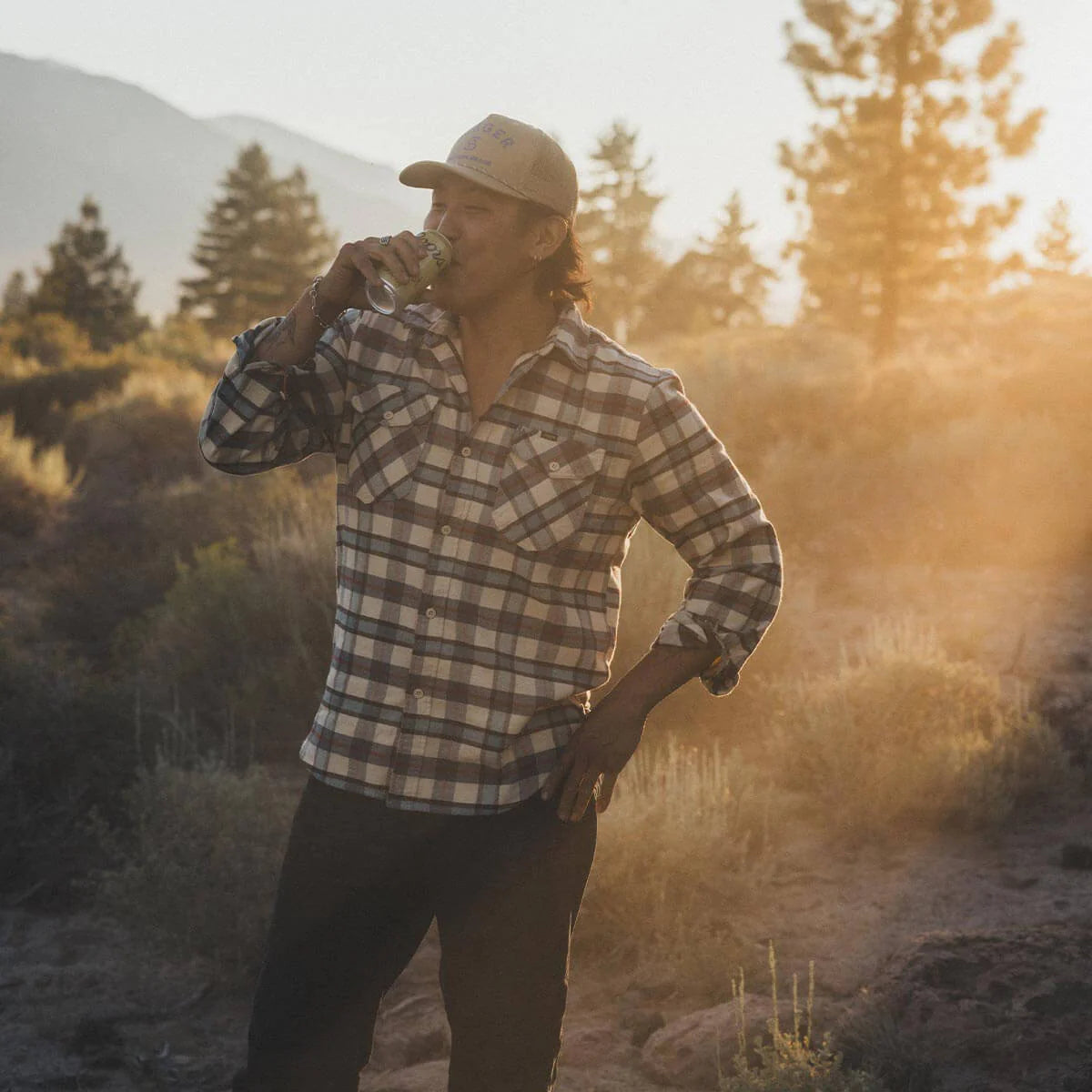 Person in a plaid shirt from Seager drinking from a bottle in a natural setting with trees and mountains.