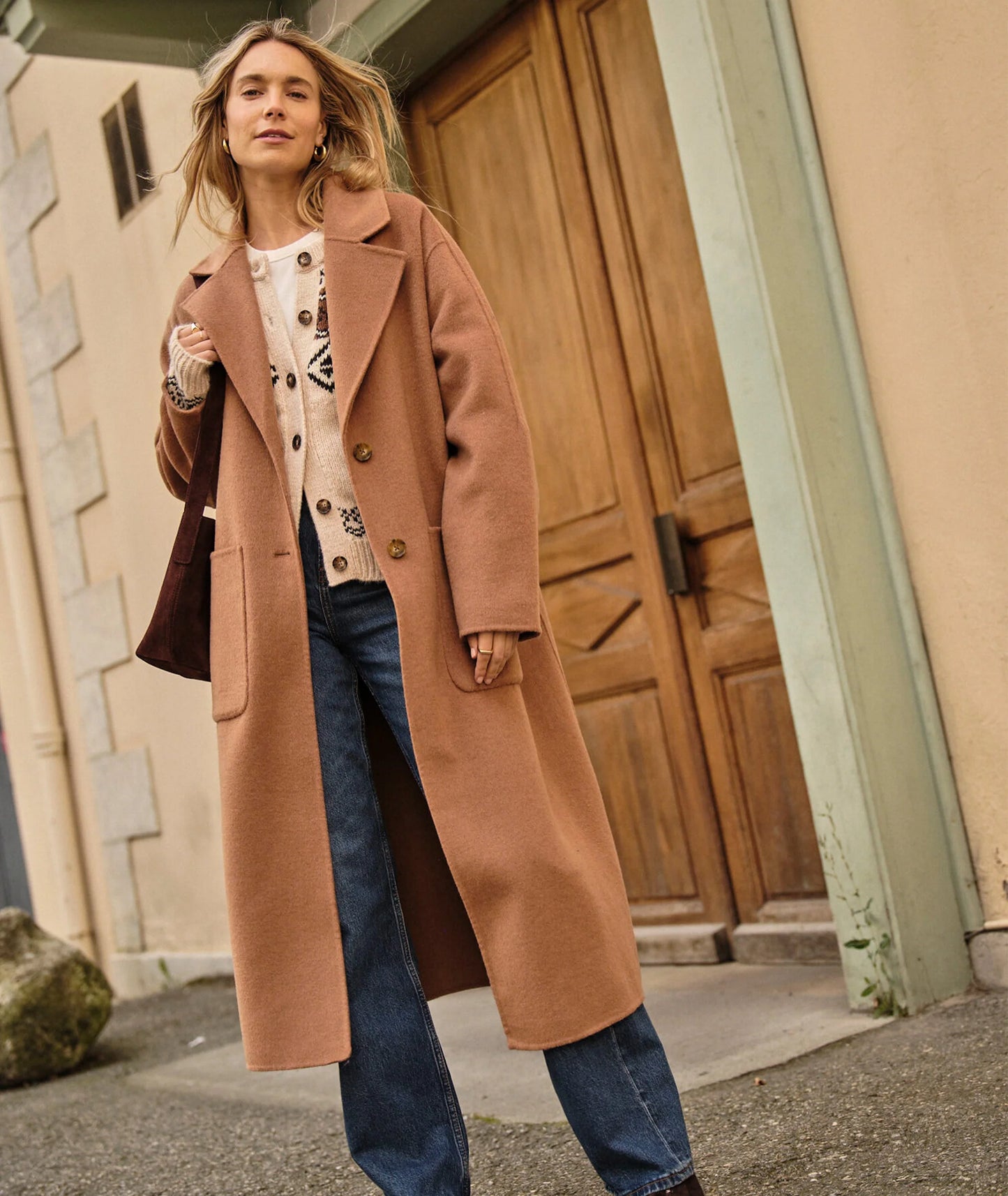 A woman wearing a camel color full-length wool coat from Marine Layer styled over a sweater and jeans.