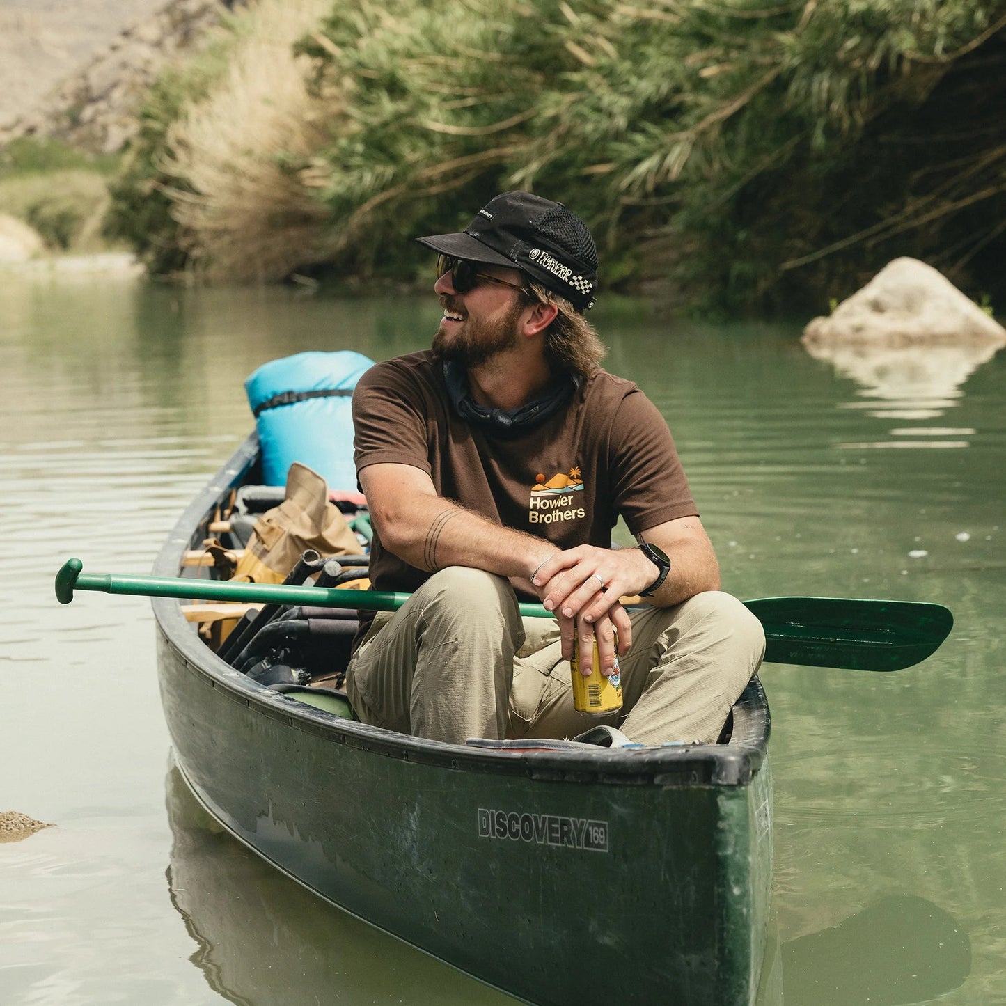 Man wearing a Howler Bros t-shirt sitting in a canoe on a calm river with trees and rocks in the background