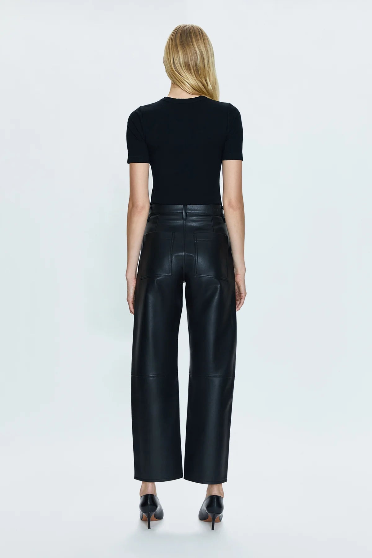 Back view of a woman wearing the Slate Black Taylor High Rise Arched Vegan Leather Trouser by the brand Pistola paired with heels and a black top