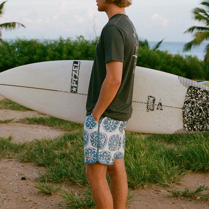 Man carrying a surfboard wearing the Howler Bros Bruja Boardshorts