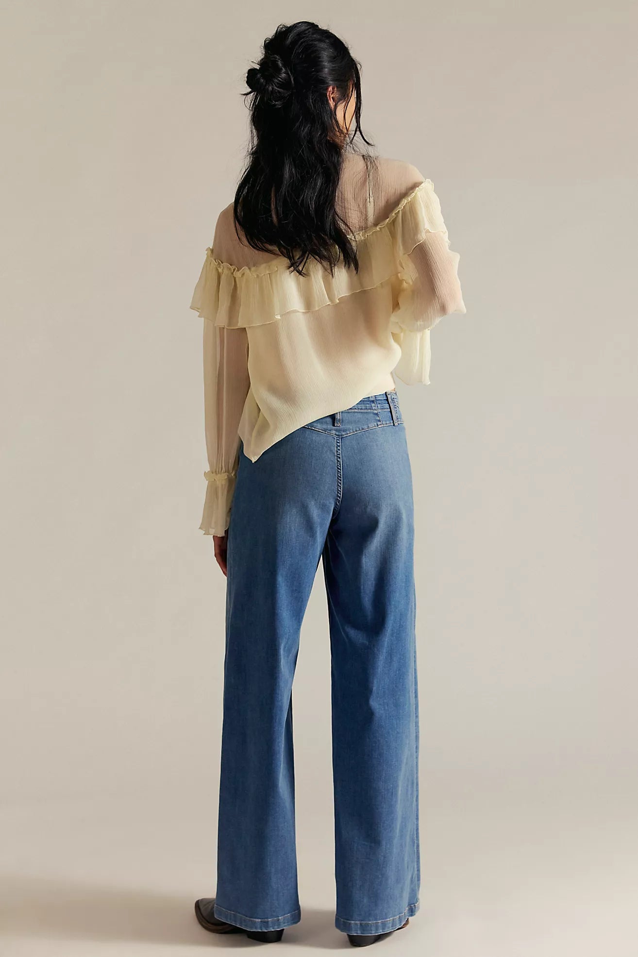 Person wearing a light yellow blouse with ruffled sleeves and blue jeans on a beige background