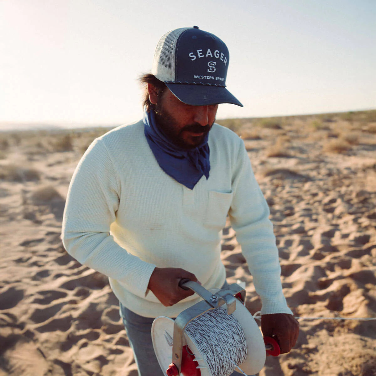 Person in a desert setting wearing a Seager cap and holding a tool.