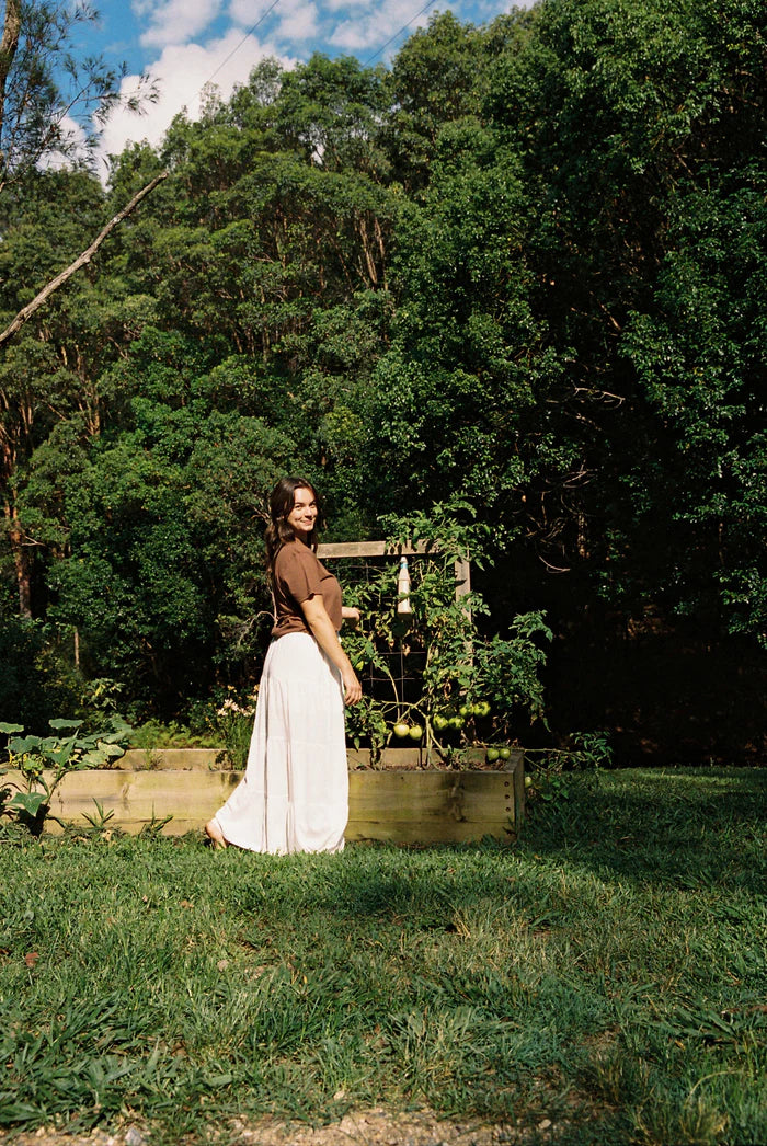 A woman outside near a garden wearing the White Classic Tiered Maxi Skirt by Rhythm