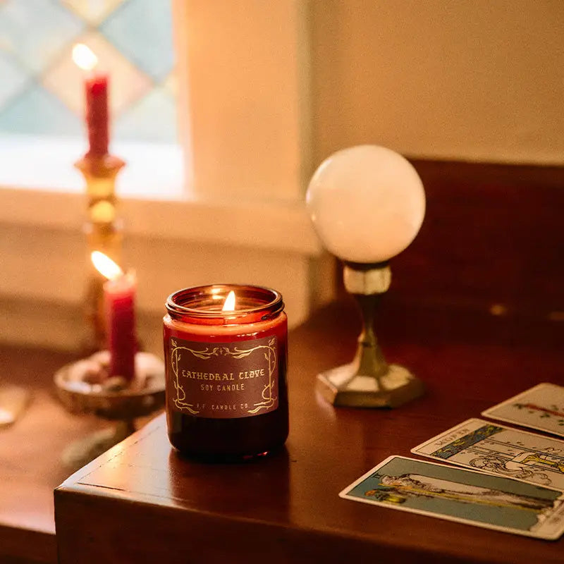 Candle with a label on a table next to tarot cards and a lamp.