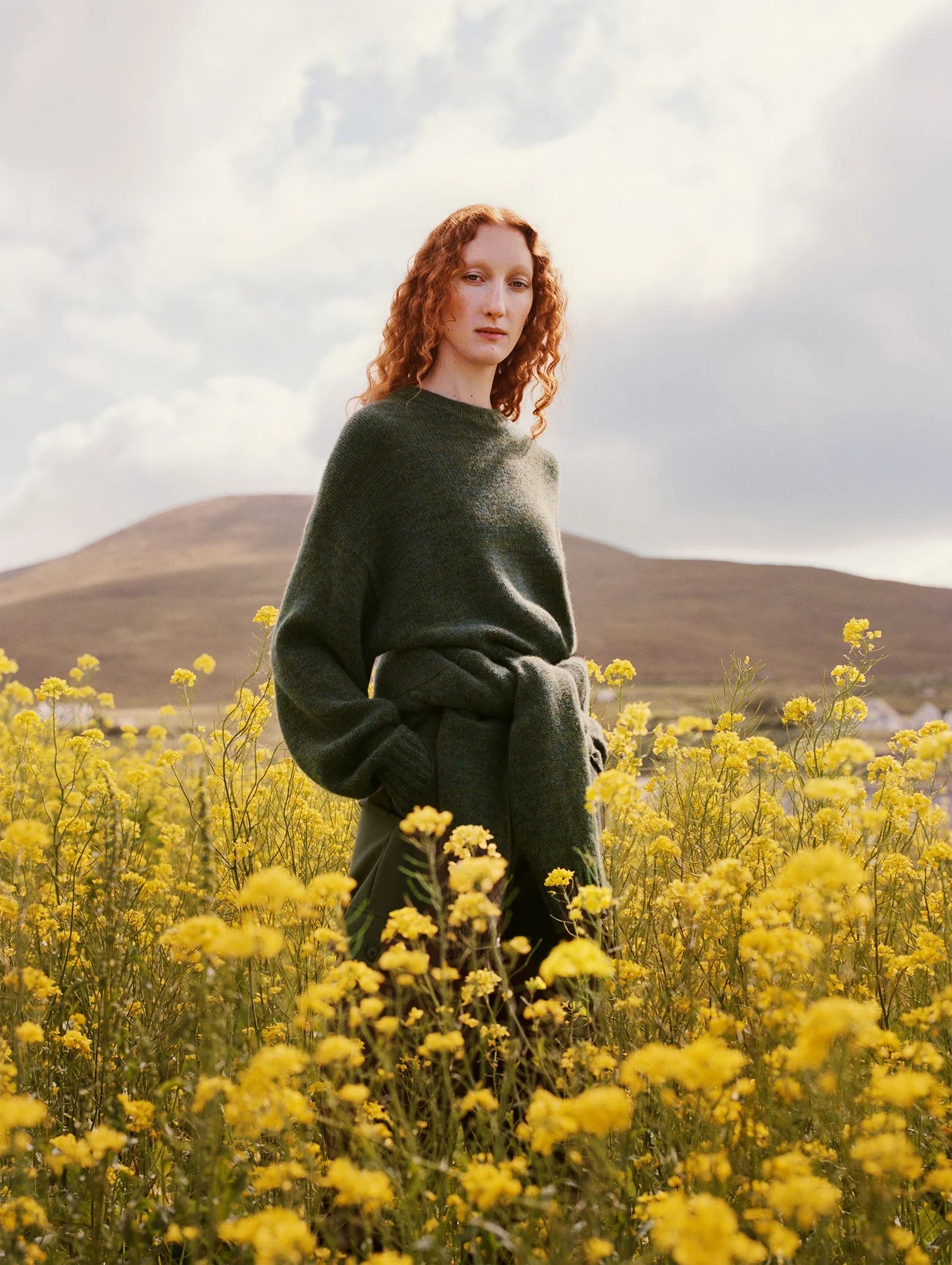 A woman standing in a field of yellow flowers while wearing the Cashmere Luxe Crewneck in the color Green Marl by the brand White + Warren