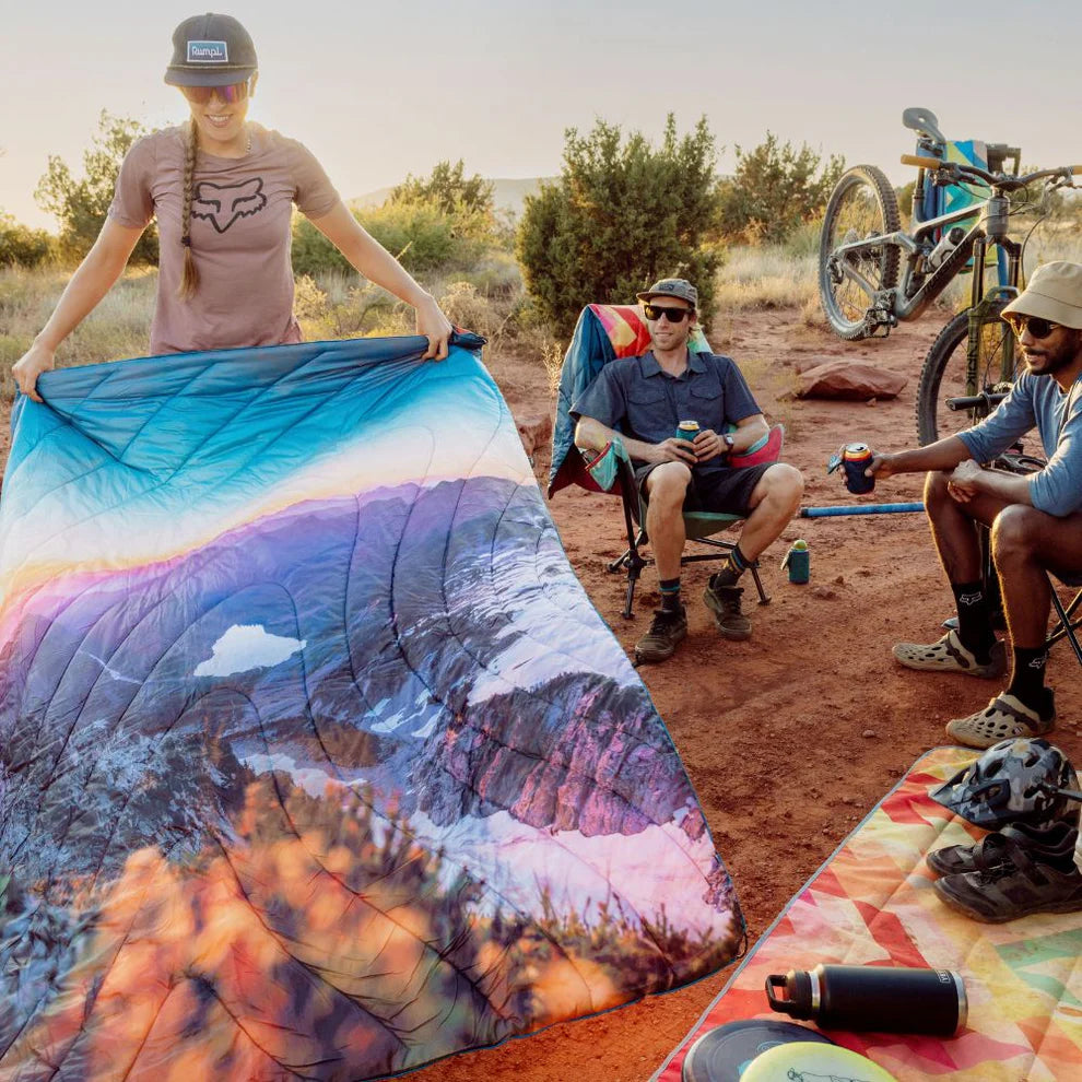 People camping outside using Rumpl's Original Puffy Blanket (1 Person) in the color Sunset Veil