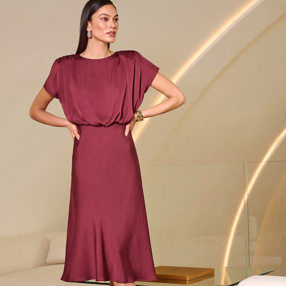 A woman wearing the Joseph Ribkoff Recycled Satin Blouson A-Line Dress while standing in a modern living room.