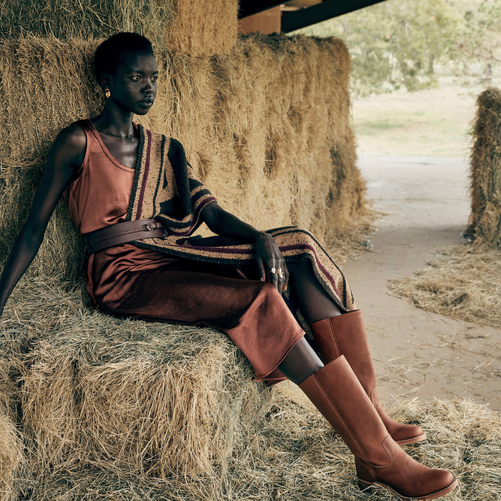 
                      
                        Woman in a rust colored slip dress sitting on hay bales in a rustic setting and wearing the Frye Campus 14L Boots in the color Saddle.
                      
                    