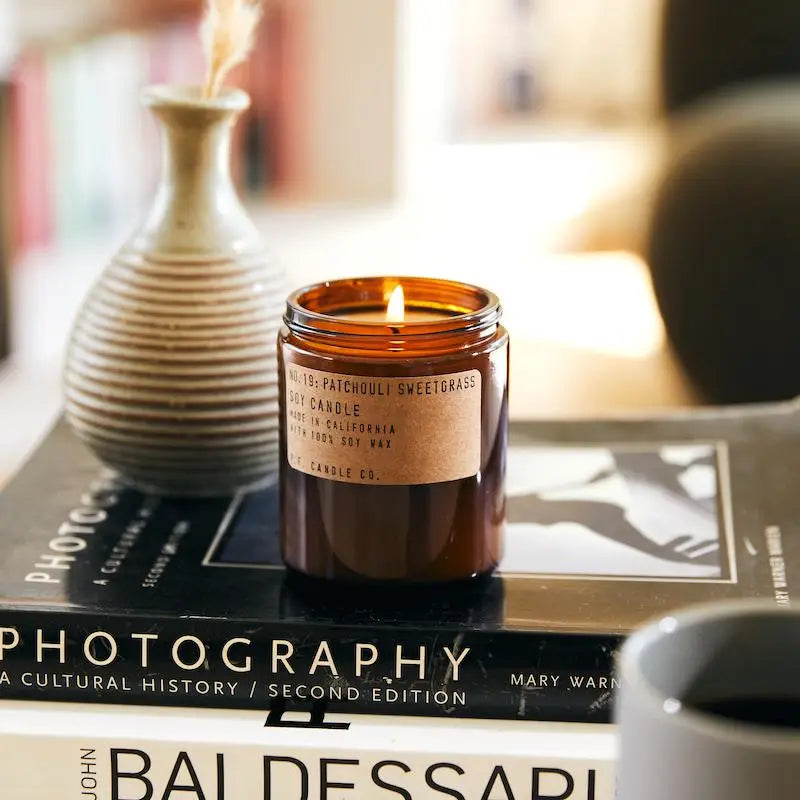 
                      
                        The Patchouli Sweetgrass 7.2 oz candle lit and posed with a decorative vase on some coffee table books
                      
                    