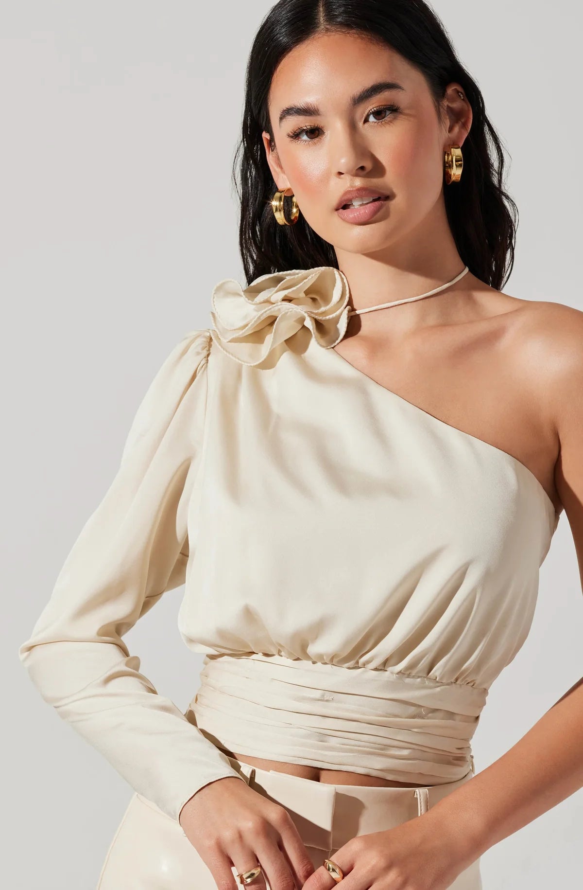 Front view of woman wearing a champagne color satin one shoulder long sleeve top with a corsage detail at the top shoulder