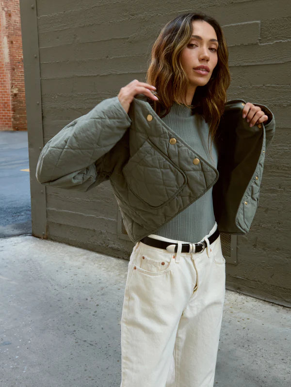 
                      
                        Woman wearing a green quilted jacket over a gray sweater, standing against a brick wall.
                      
                    