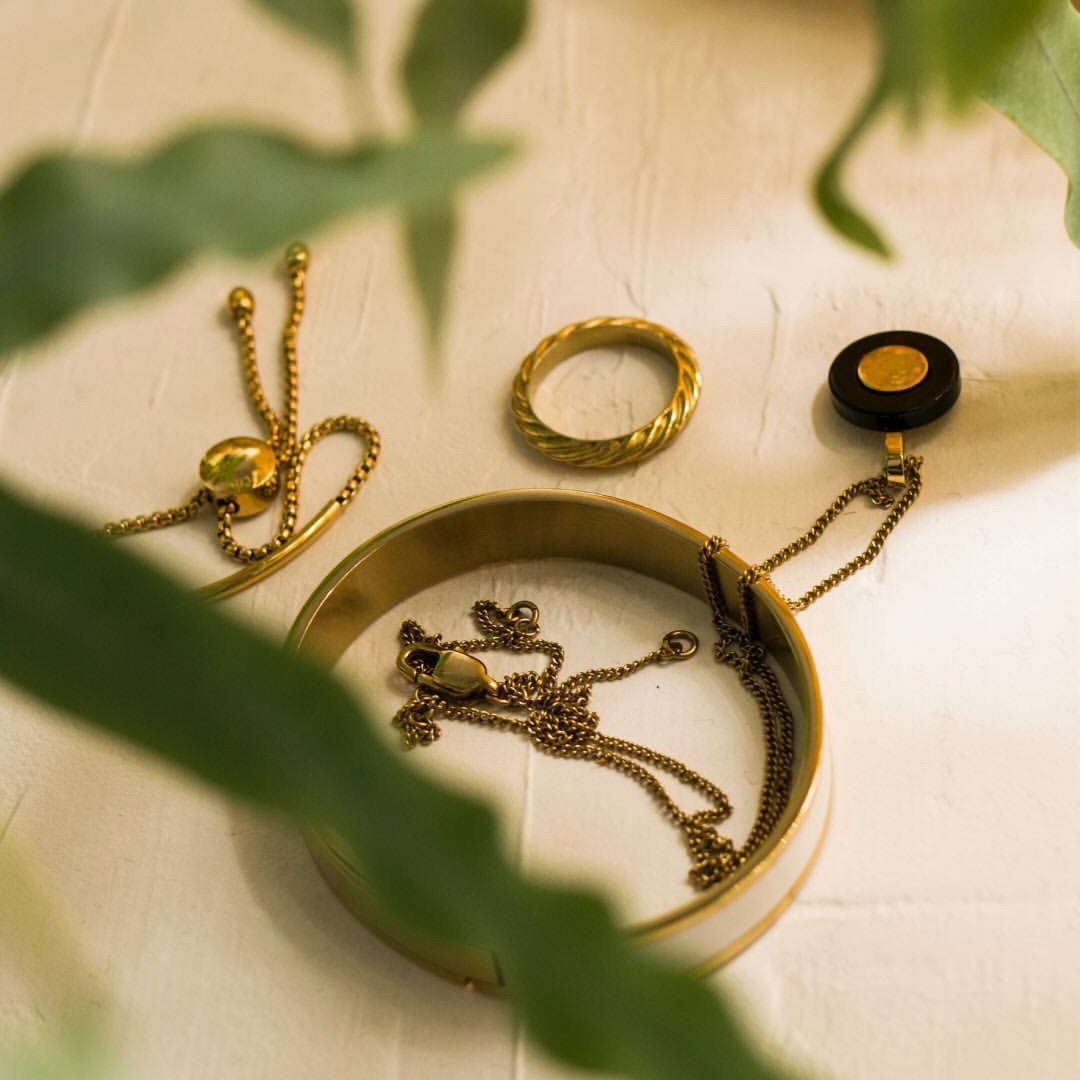 Gold jewelry including a necklace, ring, and bracelet on a textured surface with green leaves.