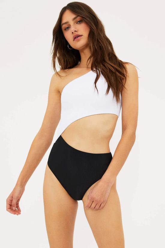 Front view of woman wearing a black and white colorblock, asymmetrical one shoulder one piece swimsuit with a side cutout detail.
