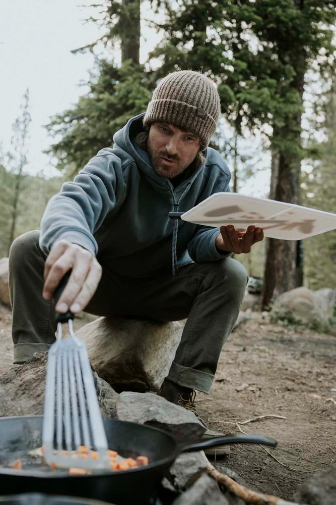 A man wearing the Bolo Fleece Hooded Sweatshirt by Roark while cooking food over a campfire in the woods