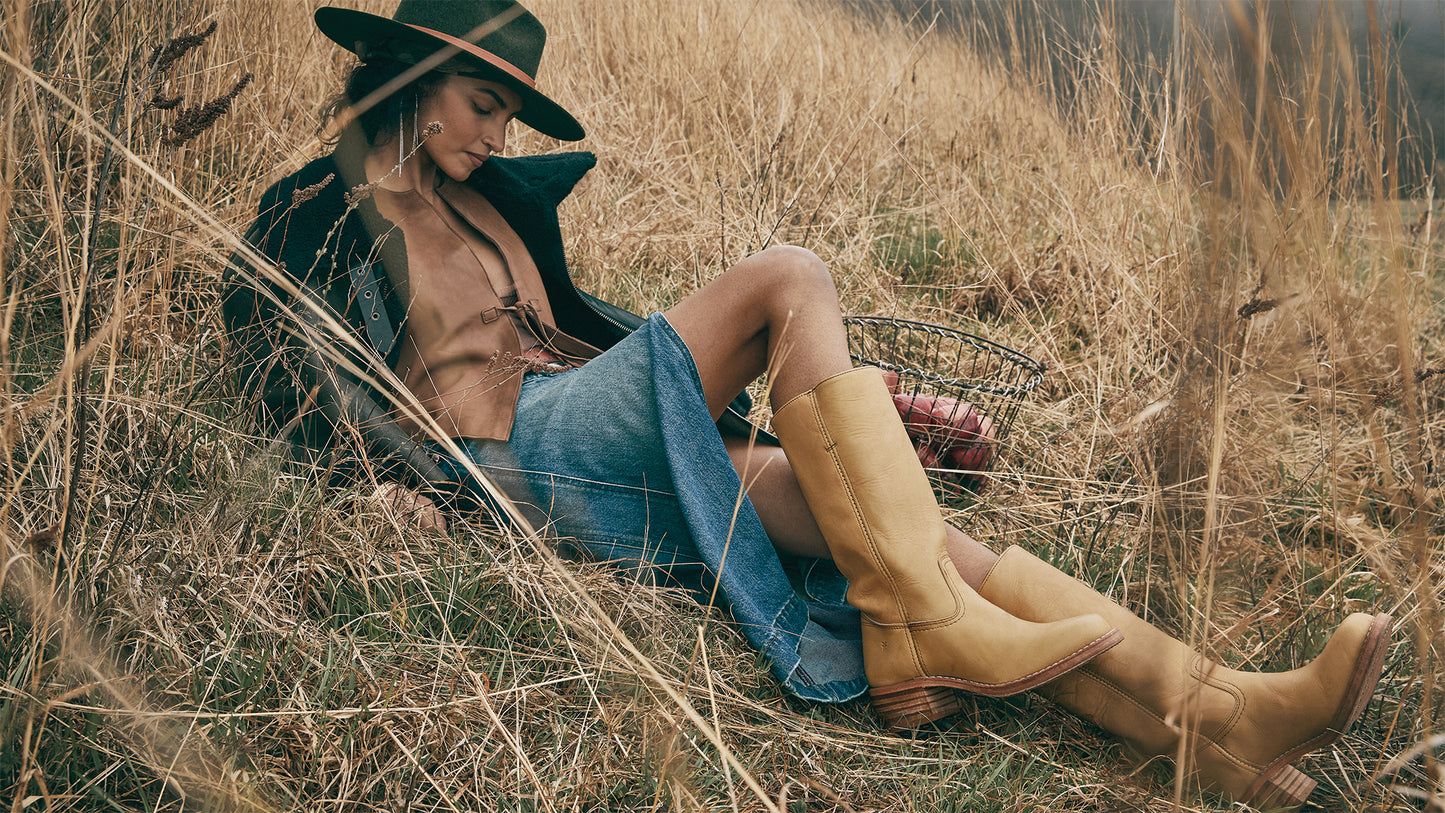 Woman is laying in tall grasses showcasing the Frye Campus 14L Boot in Banana. They are styled with a denim skirt, brown tie-front vest, open black jacket, and a black felt flat-brimmed hat.