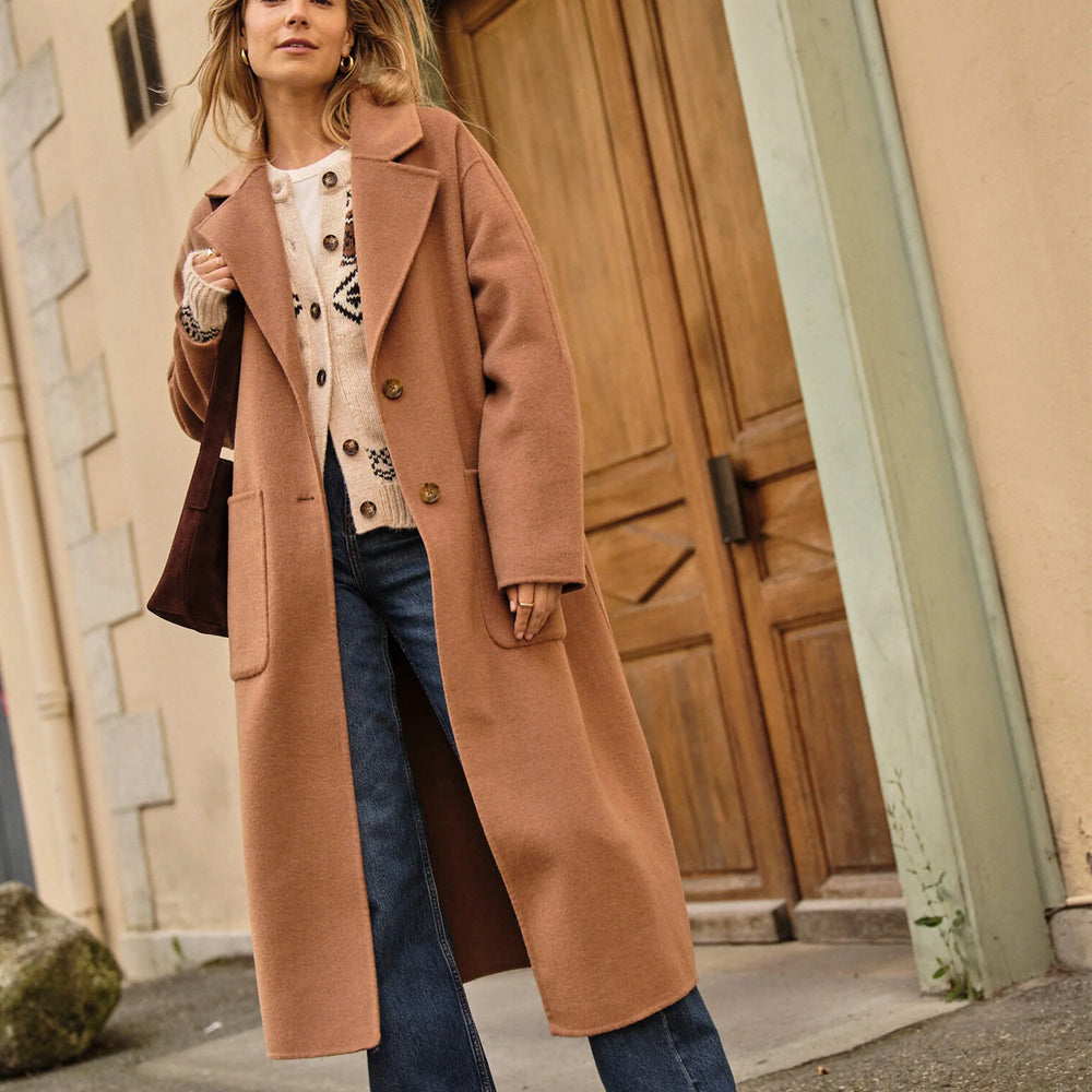 A woman wearing a camel color full-length wool coat from Marine Layer styled over a sweater and jeans. 