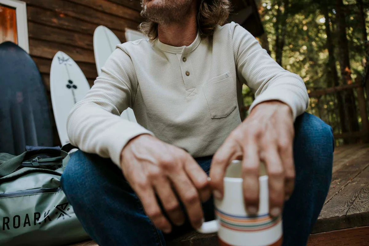 A man sitting down outside holding a coffee cup while wearing the Companion Thermal by Roark