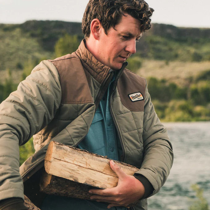 
                      
                        man wearing the green and brown Merlin Puffer Jacket by Howler Bros while carrying firewood
                      
                    