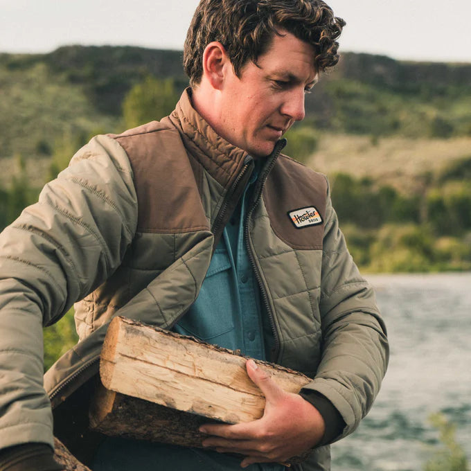 man wearing the green and brown Merlin Puffer Jacket by Howler Bros while carrying firewood