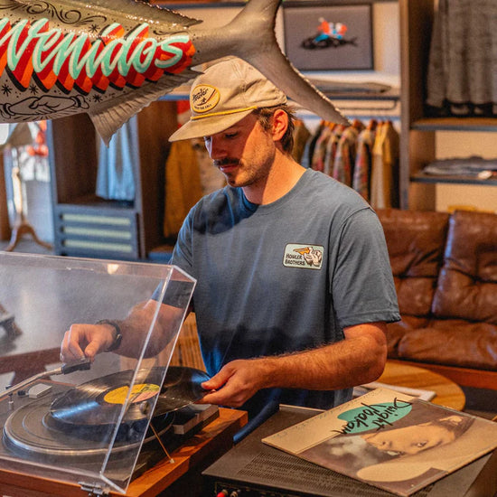 Front view of man wearing the blue Pelican Badge short sleeve t-shirt by Howler Bros while playing a record