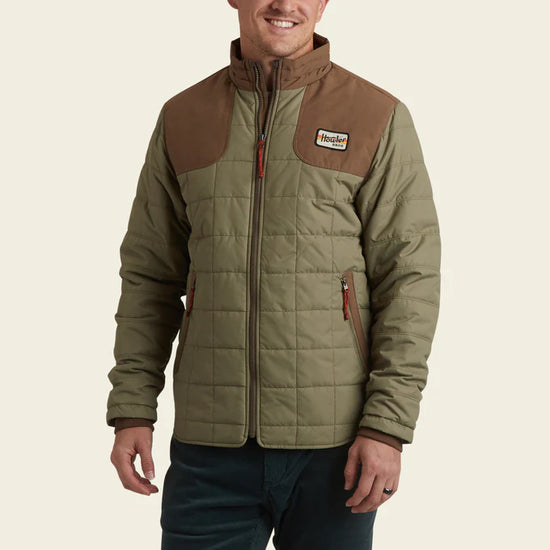 Front view of man wearing the green and brown Merlin Puffer Jacket by Howler Bros