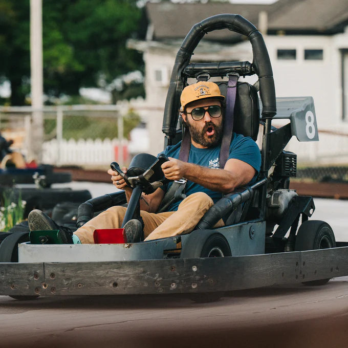 
                      
                        A man riding in a go cart while wearing the Dijon Ocean Offerings Unstructured Snapback Hat by Howler Bros
                      
                    