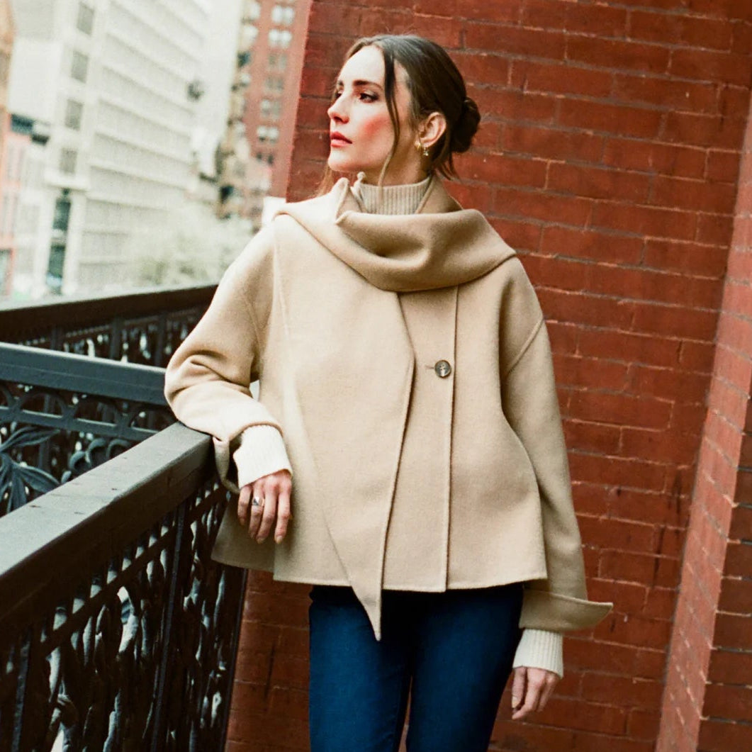 Woman in a beige coat and blue jeans standing on a balcony with a cityscape background