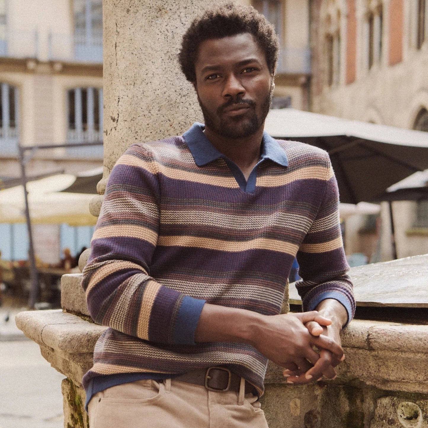Man wearing a striped sweater polo and beige pants from Marine Layer, while leaning against a stone wall