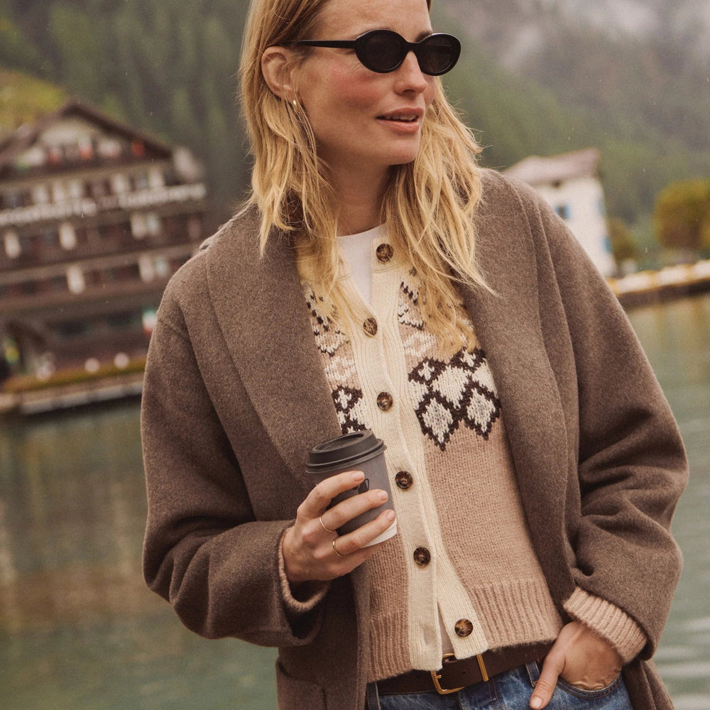 Woman wearing the Marine Layer Fiorella Cardigan layered under a brown coat. She's holding a coffee cup, standing by a lake with mountains in the background.