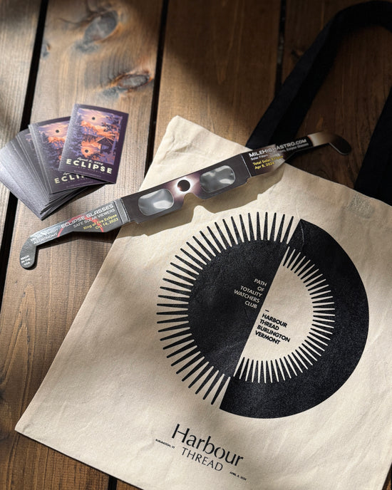 Flat lay view of the custom Harbour Thread Eclipse Tote Bag, eclipse glasses, and eclipse sticker