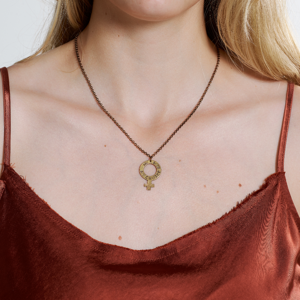 
                      
                        Woman wearing a brass necklace with a pendant, wearing a rust-colored top.
                      
                    