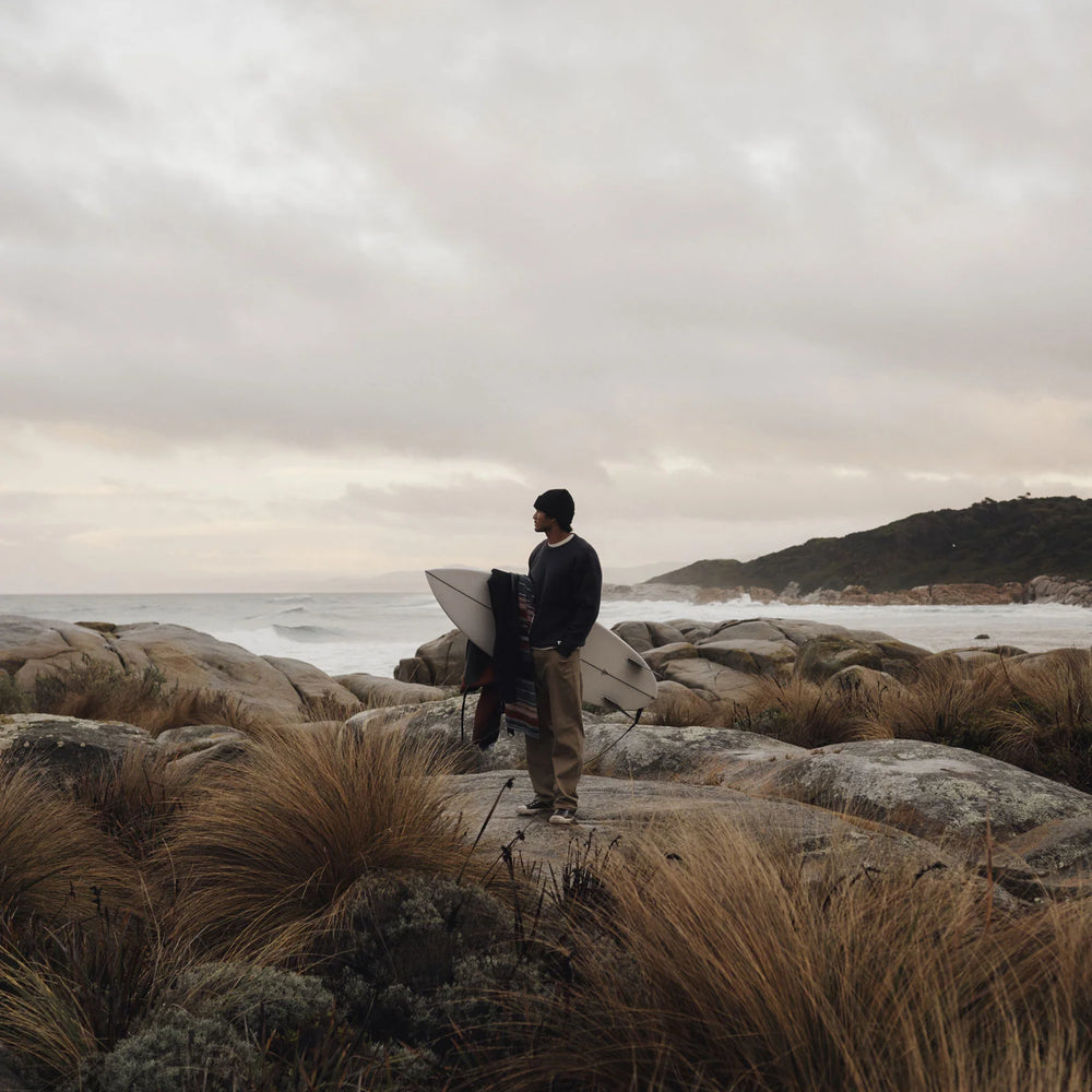 Man wearing the Rivers Sweater by Katin while holding a surfboard on a rocky beach with a cloudy sky