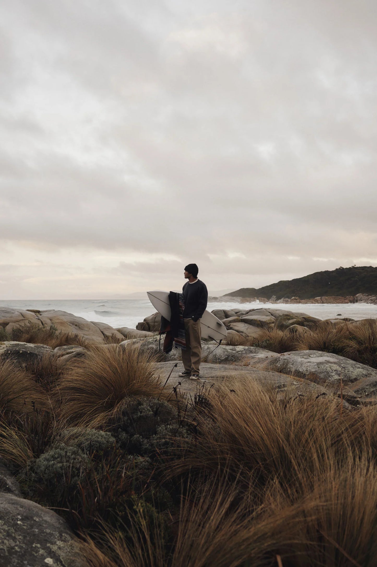 Man wearing the Rivers Sweater by Katin while holding a surfboard on a rocky beach with a cloudy sky