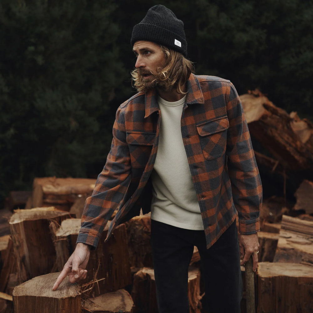 Man in a plaid shirt and beanie from Katin standing among stacked wooden logs in a forest setting