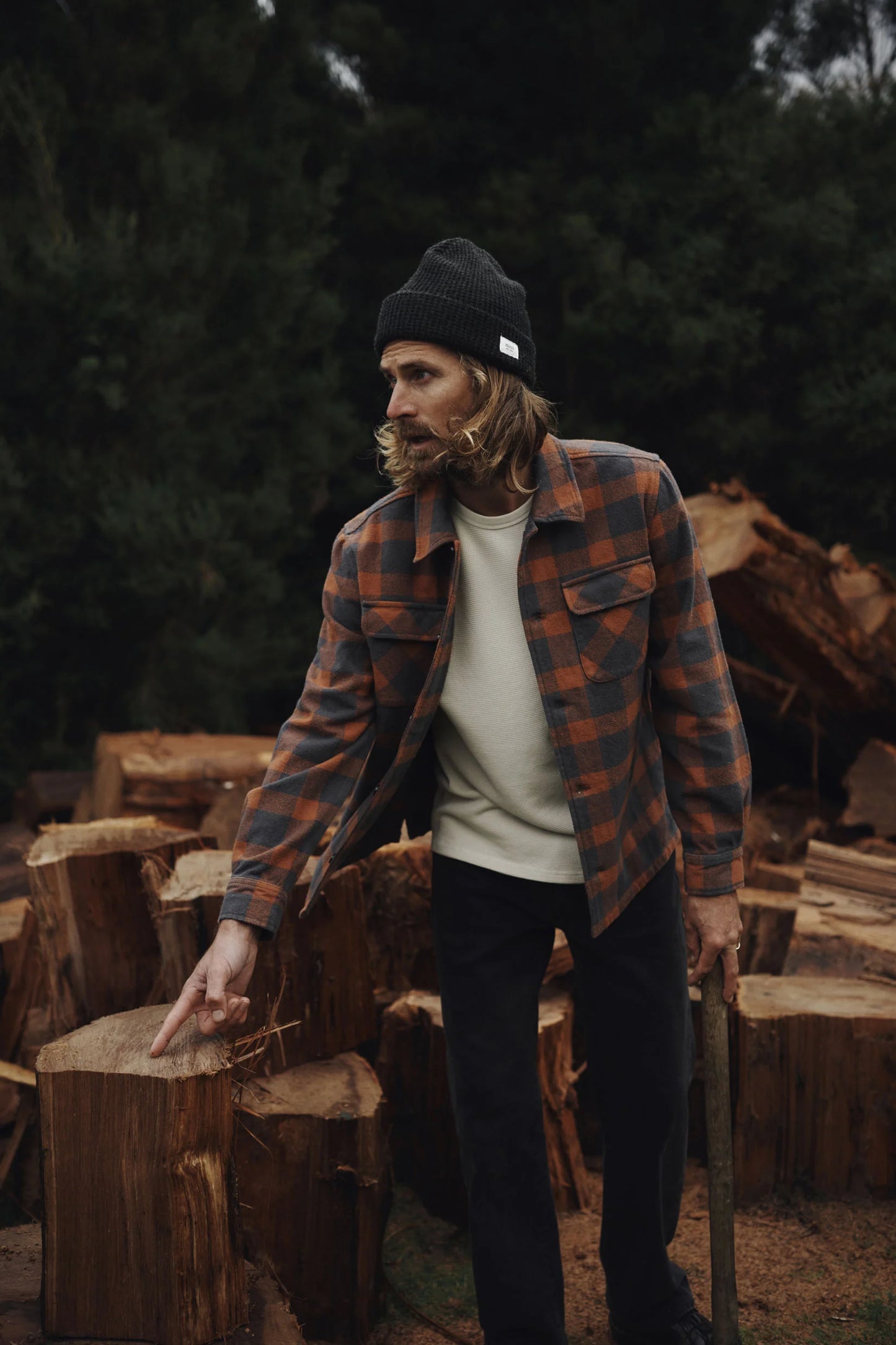 Man in a plaid shirt and beanie from Katin standing among stacked wooden logs in a forest setting