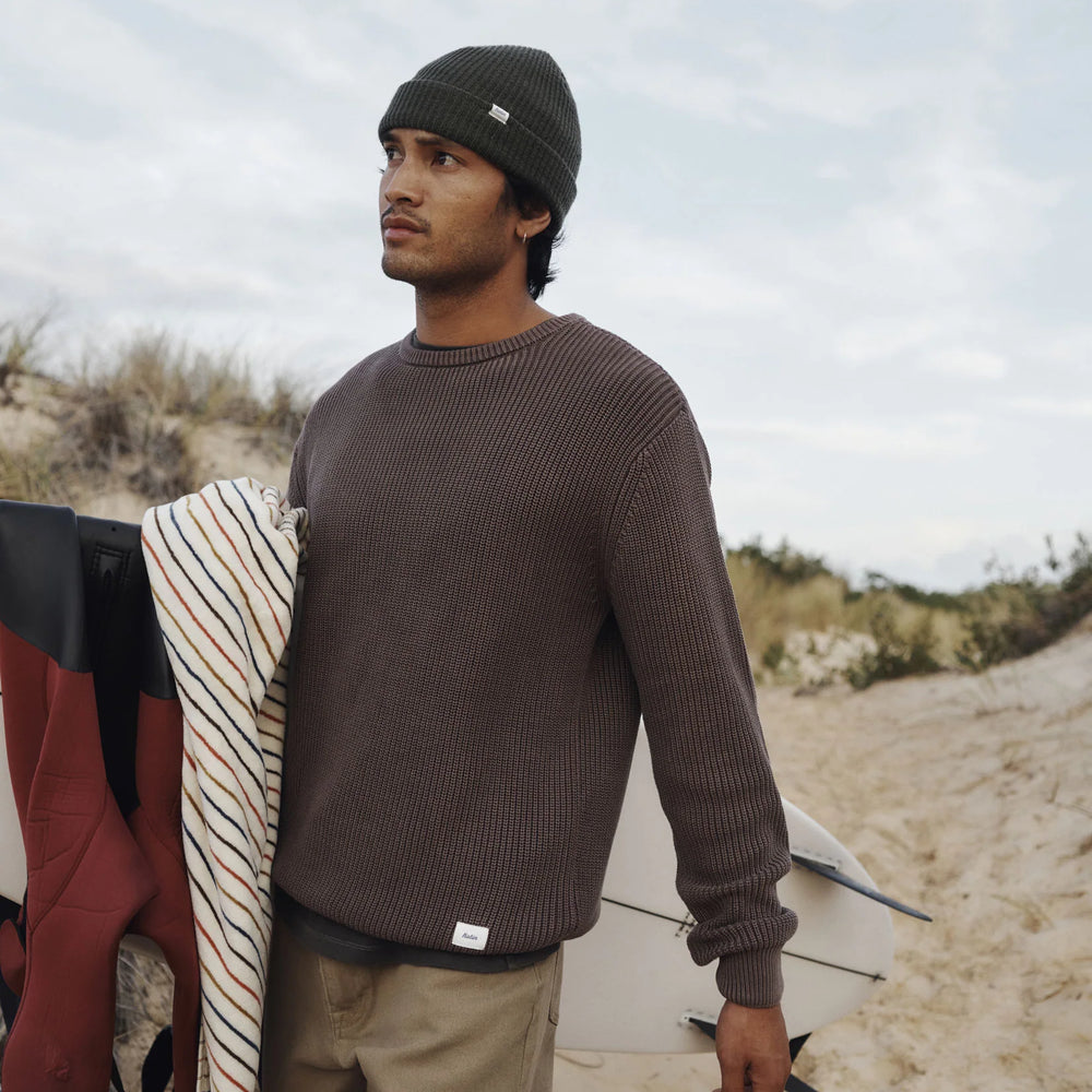 Man walking on a sandy path with a surfboard, wearing a beanie and sweater from Katin