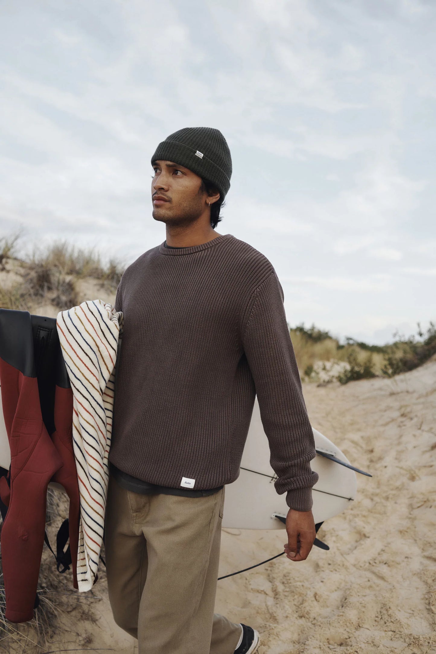 Man walking on a sandy path with a surfboard, wearing a beanie and sweater from Katin