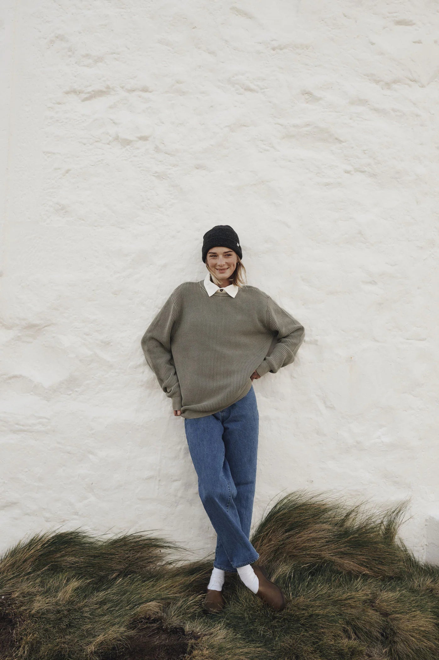 Woman wearing the Moss Swell Sweater by Katin paired with blue jeans standing against a white textured wall with grass at the bottom.