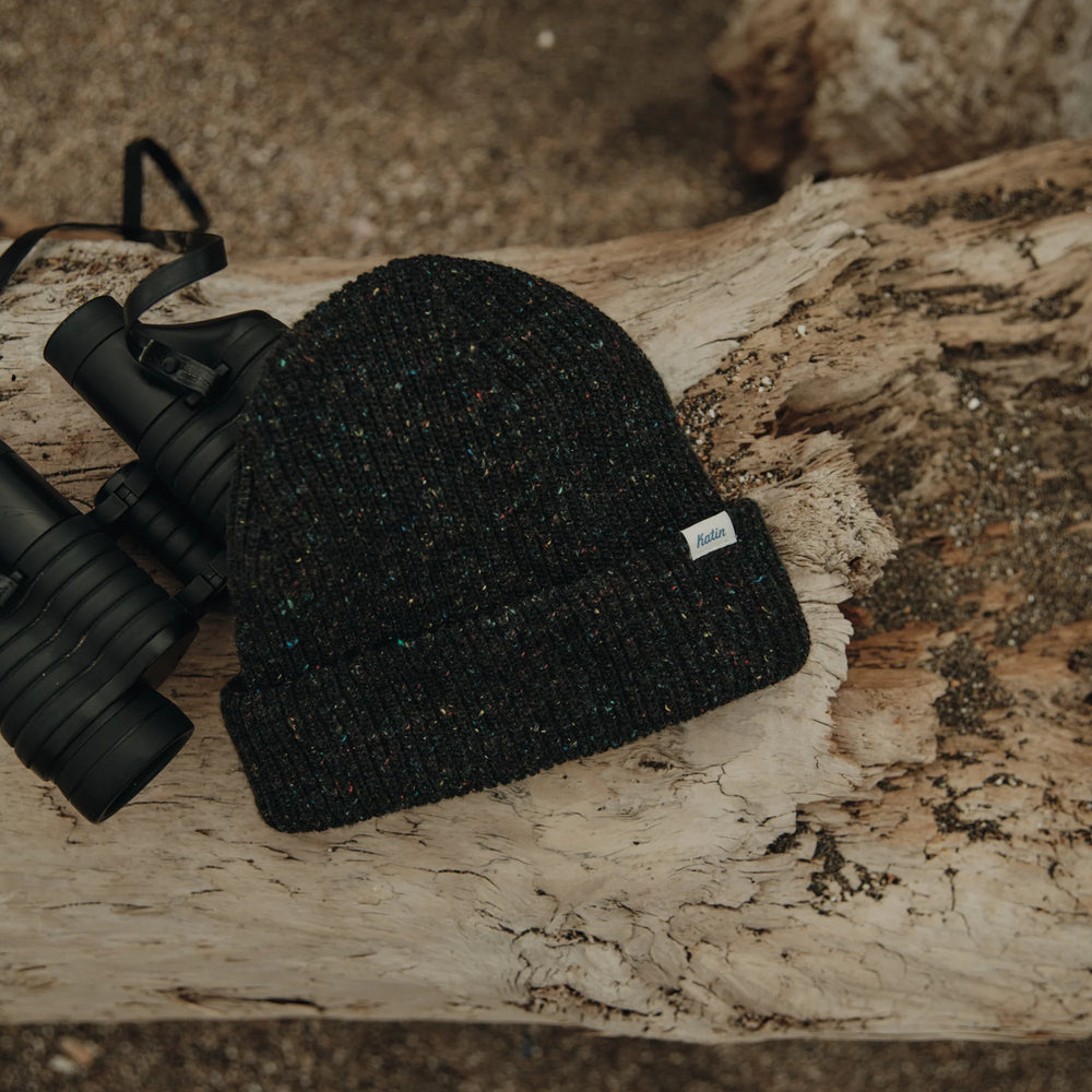 Black knit beanie from Katin next to binoculars on a wooden log