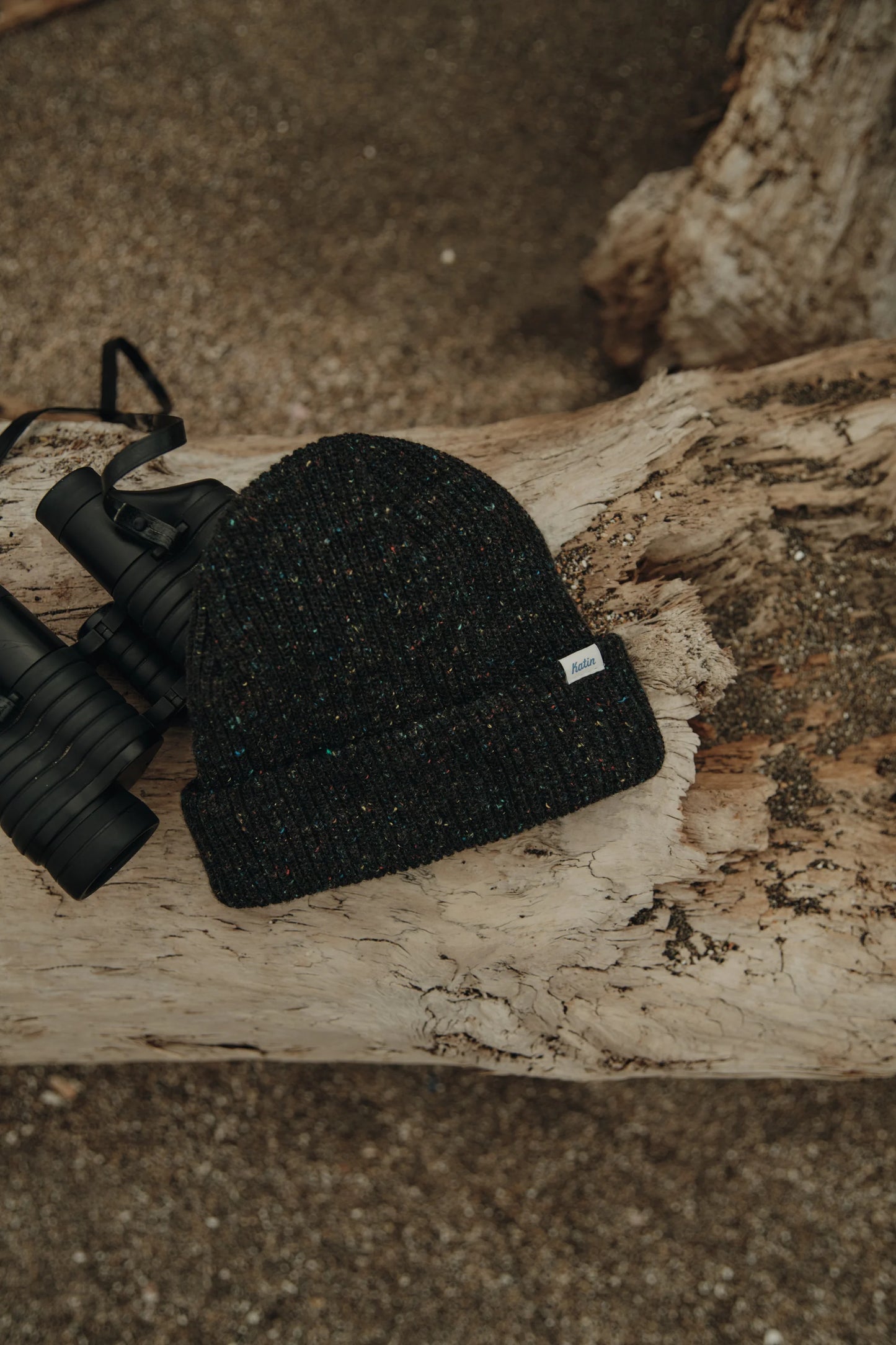 Black knit beanie from Katin next to binoculars on a wooden log