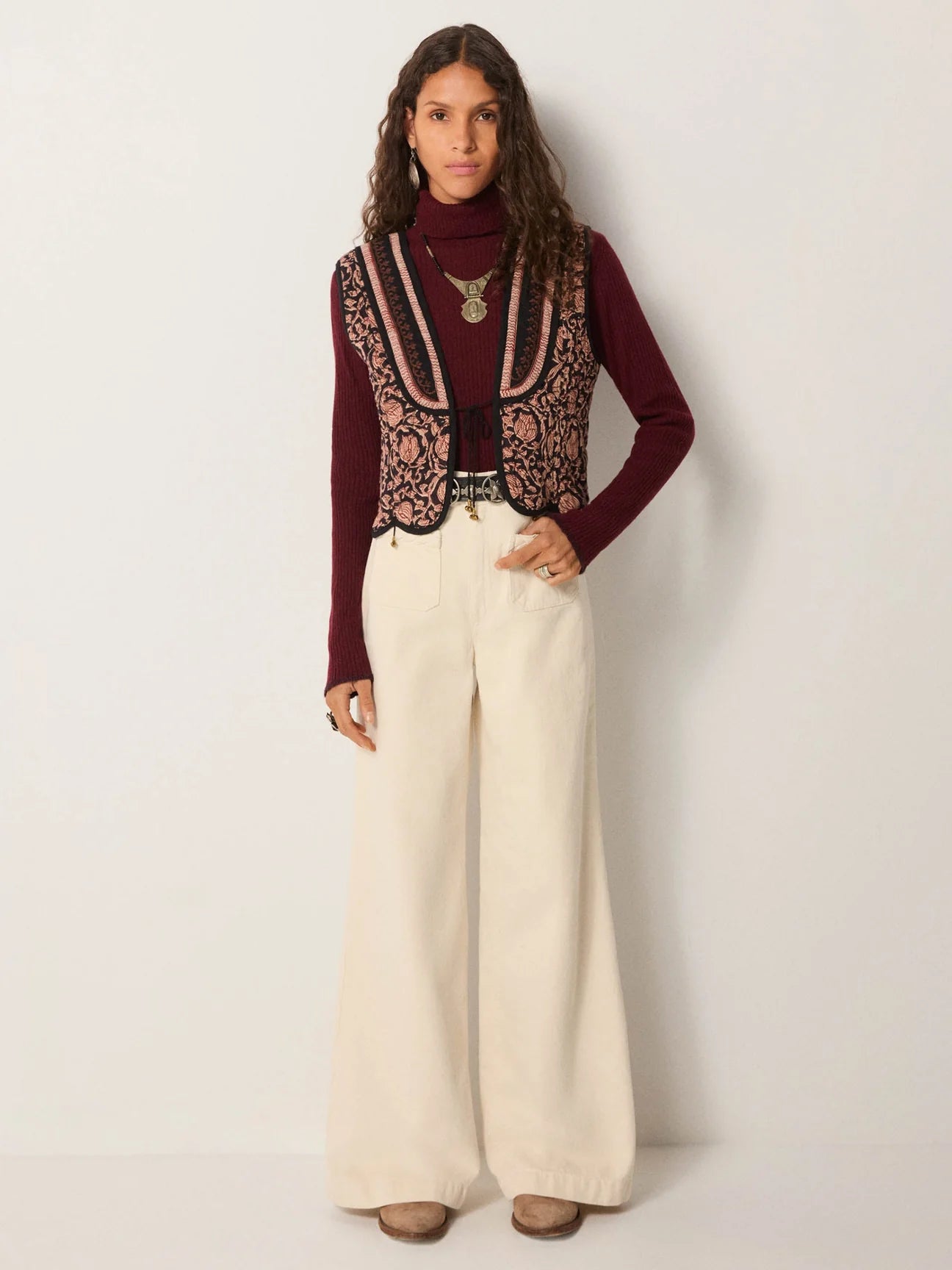 Person wearing a burgundy sweater, patterned vest, and white pants from Maison Hotel