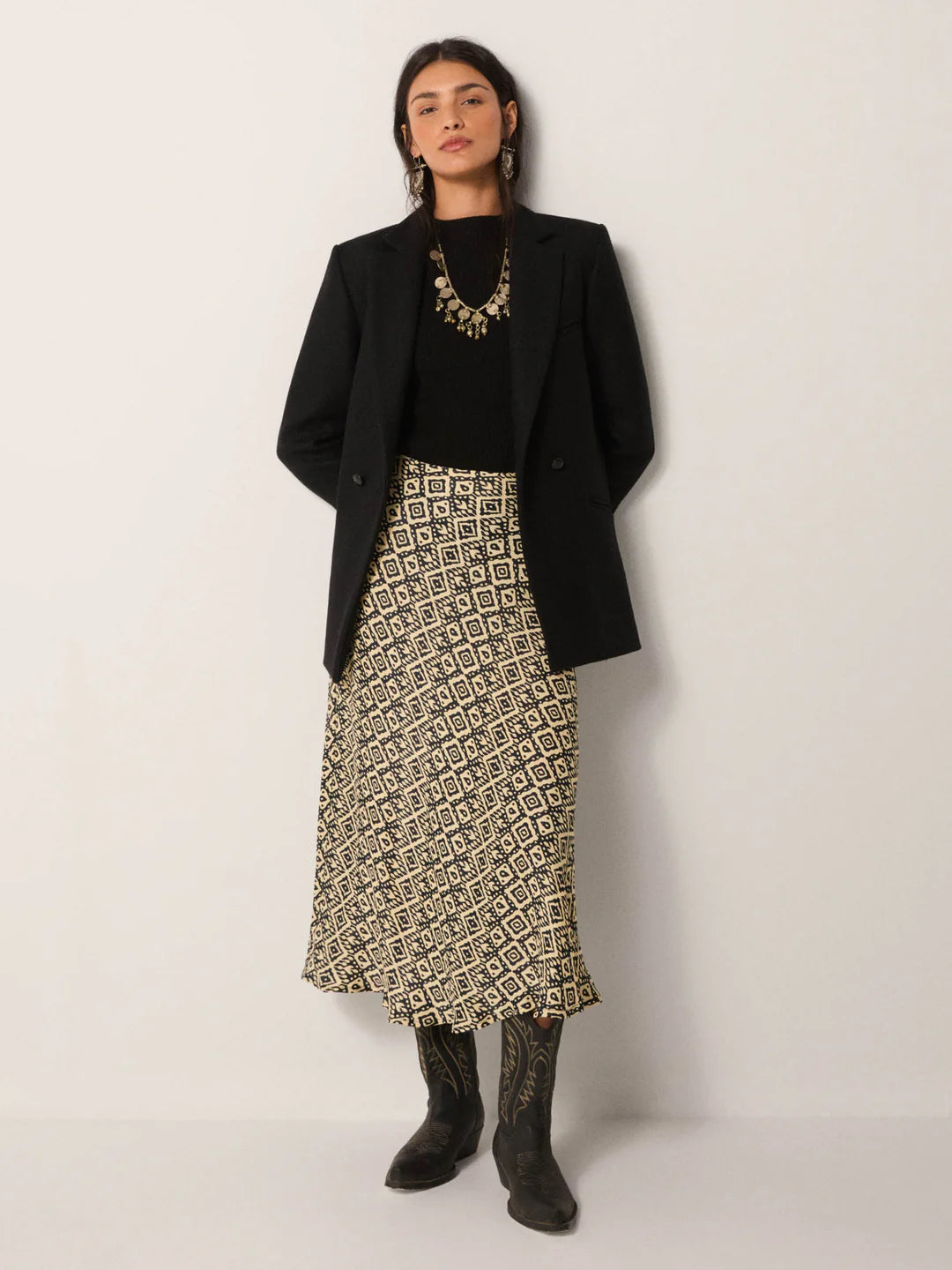 A woman wearing the Irene Skirt from Maison Hotel paired with a black top, black blazer, black cowboy boots and a statement necklace