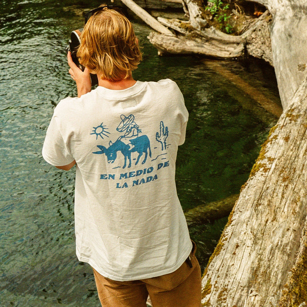 
                      
                        A man standing on a log while taking a photo of water on an old camera. He's wearing the white short sleeve Nowhere Tee from the brand Seager.
                      
                    