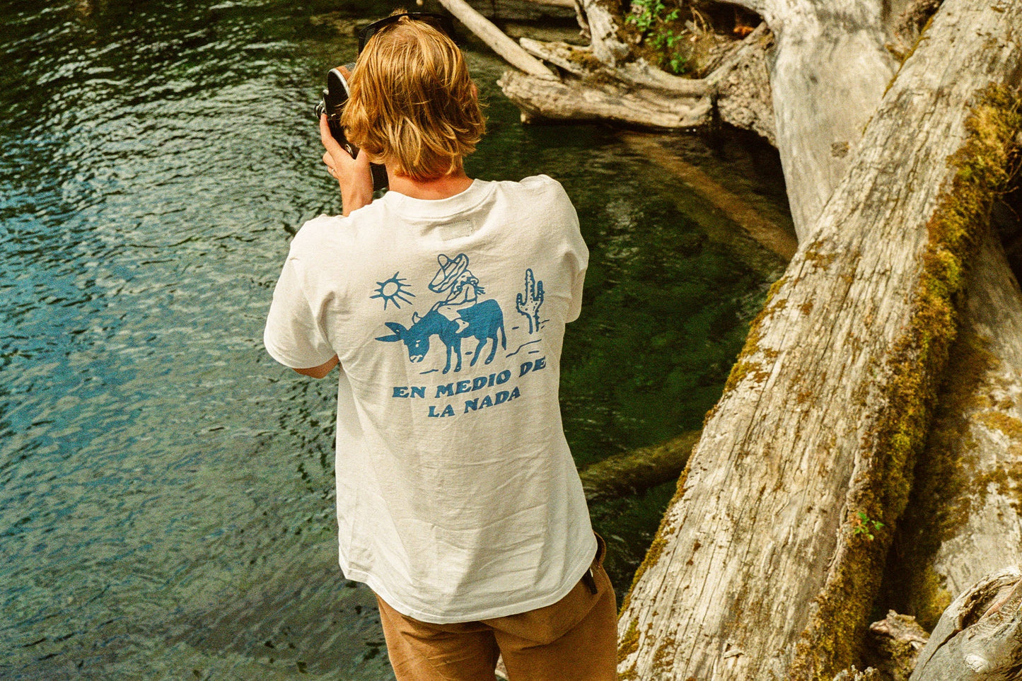 A man standing on a log while taking a photo of water on an old camera. He's wearing the white short sleeve Nowhere Tee from the brand Seager.