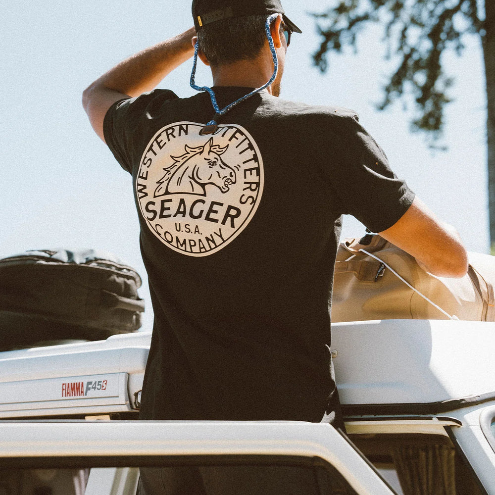 
                      
                        A man standing outside looking over the top of a car. He's wearing the black short sleeve Stang Tee by the brand Seager. 
                      
                    