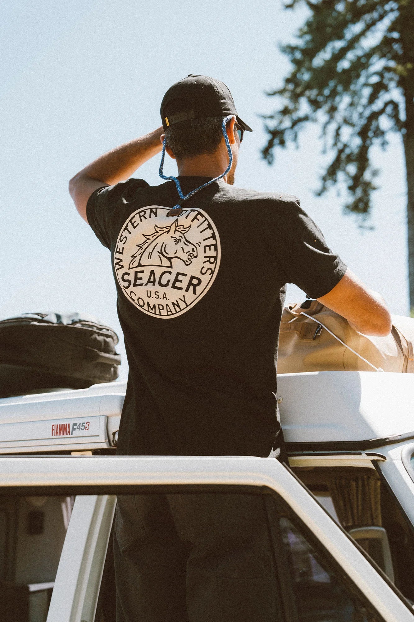 A man standing outside looking over the top of a car. He's wearing the black short sleeve Stang Tee by the brand Seager. 